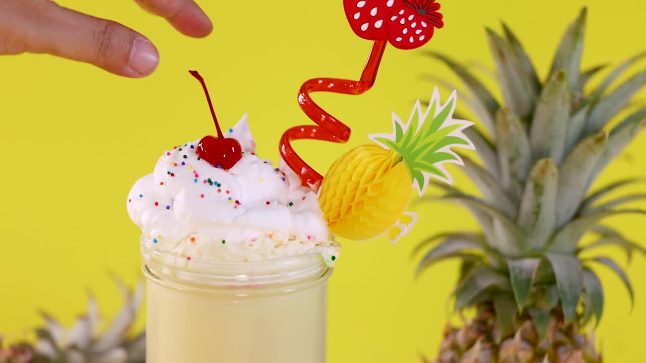 Hand lifts cherry from whipped cream-topped pineapple smoothie, colorful straws, bright yellow background, static shot