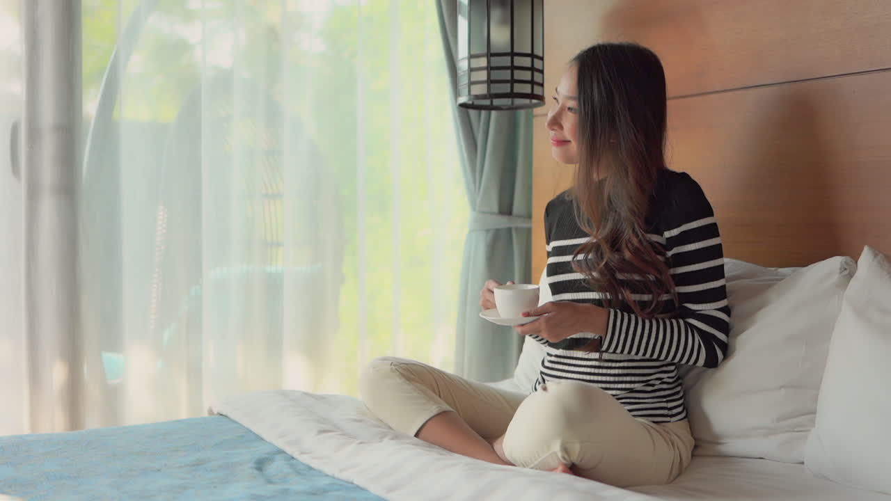 A woman sitting up in bed while drinking a cup of coffee turns to look out the window