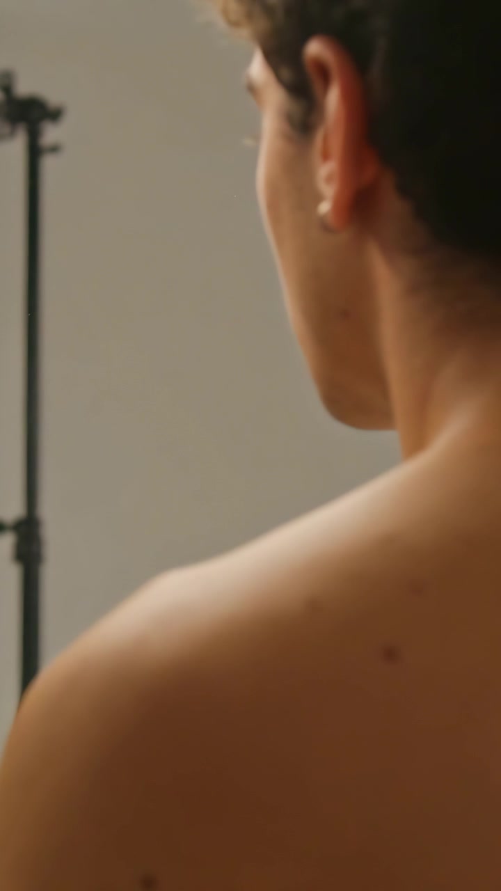 Vertical video: Looking at mirror, shirtless teen adjusting hair for shoot, earring and light stand