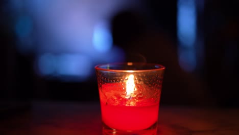 red candle at night burning, flickering in glass jar on marble table with large tv television on and people moving around in background silhouette - in 4k 30fps