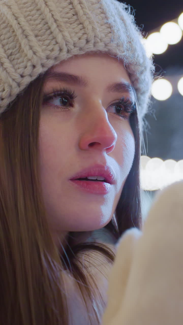 Young girl sits outdoors in winter, covering her face with gloved hands before exhaling visible cold breath, city lights glow behind her as people move around in a lively, festive urban environment