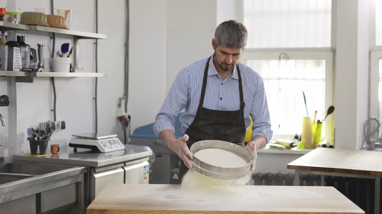 красивый мужчина пекарь просеивает муку через сито на кухне.