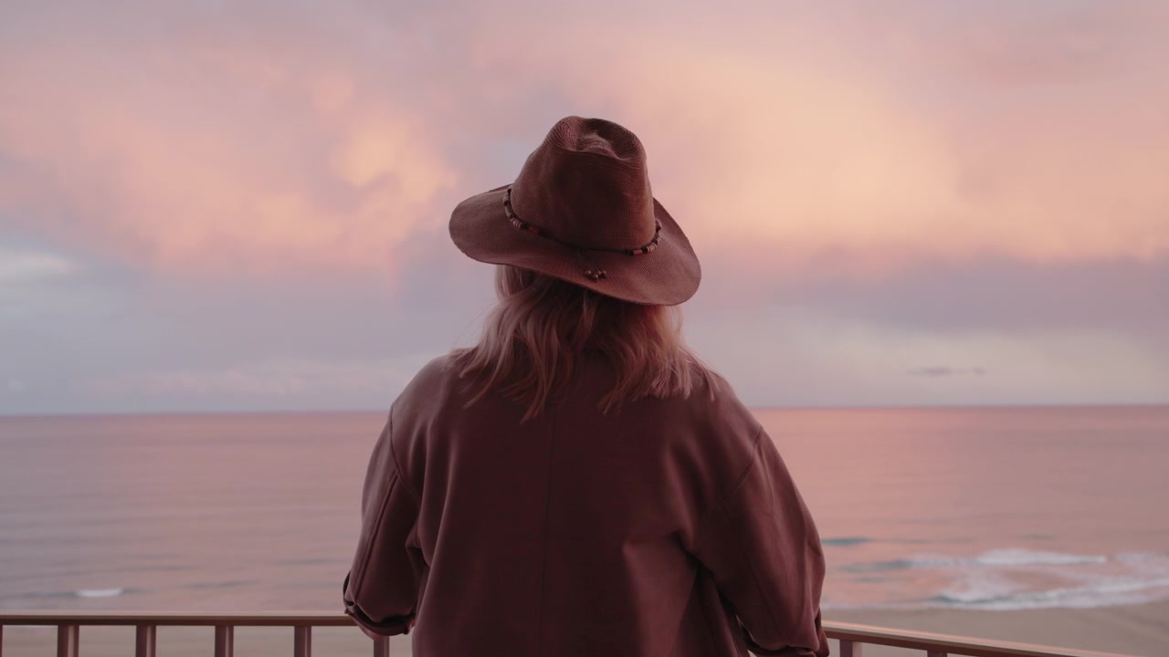 Kvinde med hat ser solnedgang over havet fra balkon