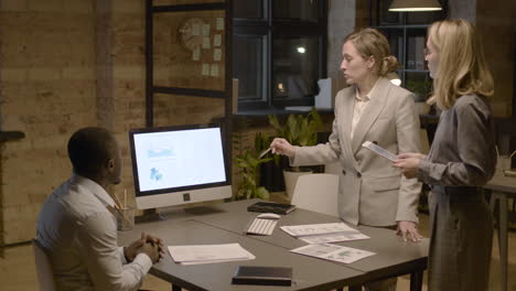 american man sitting at desk in the office while talking about graphics and stadistics with two female coworkers who are standing 1