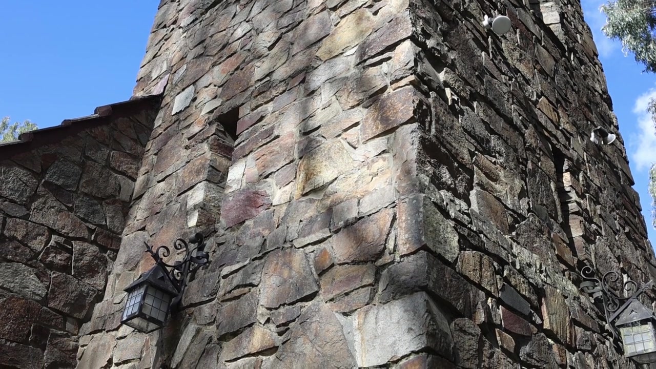 Filmagem em grande angular de uma rústica torre de igreja de pedra antiga em Olinda, Melbourne. A luz do sol projeta sombras na pedra texturizada contra um céu azul vívido, enfatizando a arquitetura clássica. Nenhuma pessoa visível.