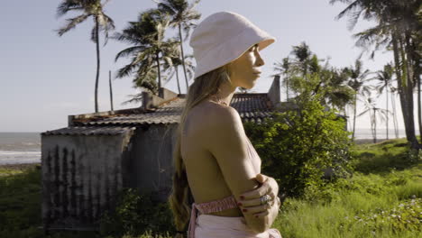 Blonde-woman-with-braid-and-crossed-arms-near-abandoned-house-on-beach