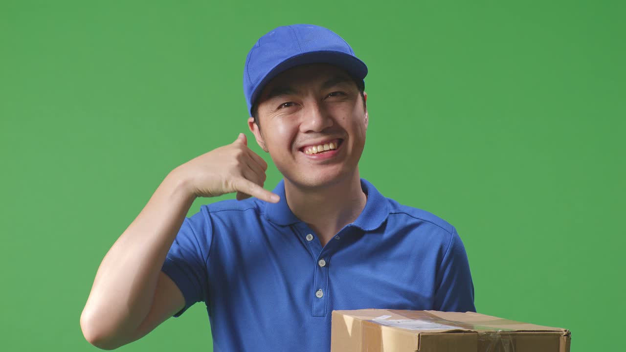 푸른 유니폼을 입은 아시아 남성 택배사의 클로즈업은 스튜디오에서 녹색 스크린 배경에 상자를 배달하는 동안 제스처와 미소를 지으며 나를 호출합니다.