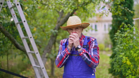 male gardener in hat holding a fresh ripe apple outdoors. farmer sniffs aromatic apple in the garden in autumn. organic food.