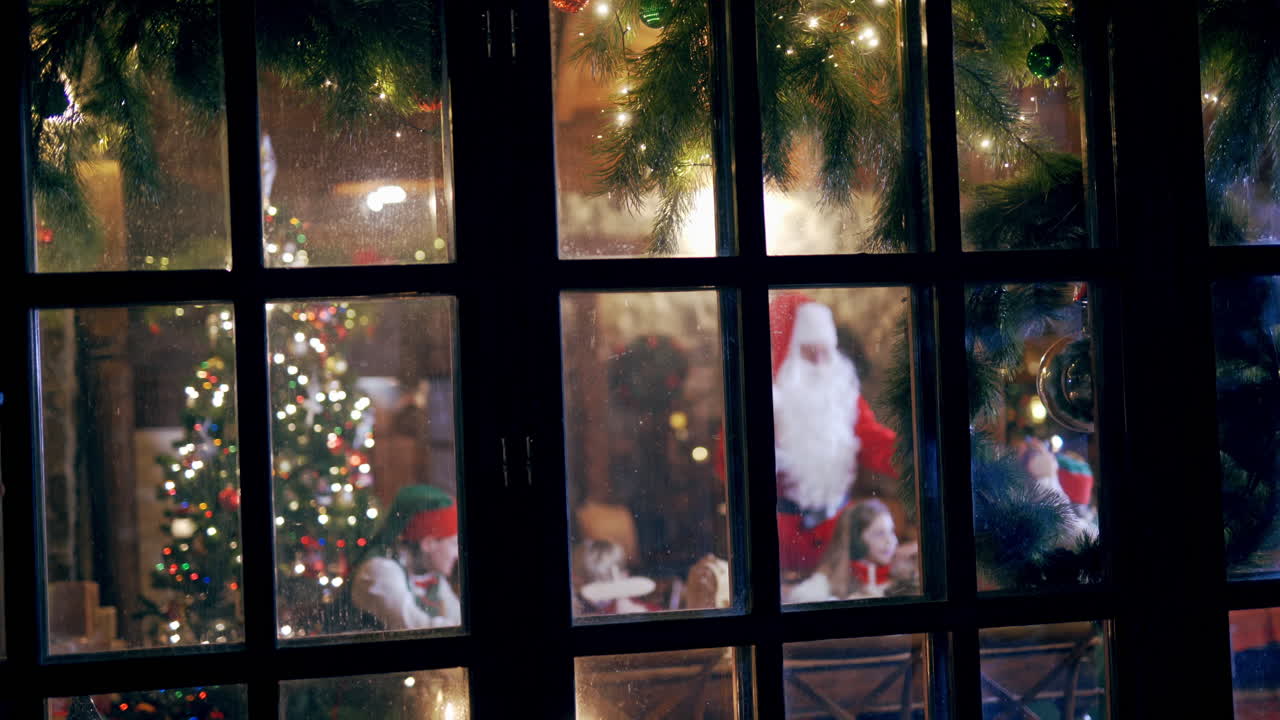Joyful atmosphere with Christmas decorations inside the house. View from the street into the cozy house with children and Santa having a good time together in the evening.