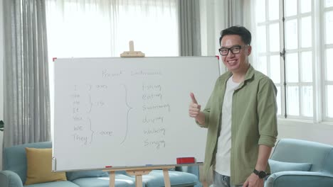 asian male teacher with glasses showing thumbs up gesture and smiling to camera while teaching english at home