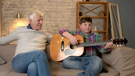 the grandson plays for his grandfather guitar, music. an elderly man is sitting on with a young fat guy playing guitar. home comfort, family idyll, cosiness concept, difference of generations 60 fps