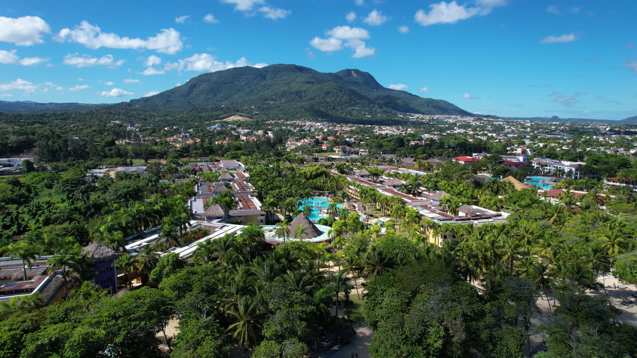 Aerial approaching shot of Costa Dorada Beach and luxury resort hotel in Puerto Plata, Dominican Republic. Sunny day on tropical island with sunshades and palm trees. Clear turquoise water of Bay.