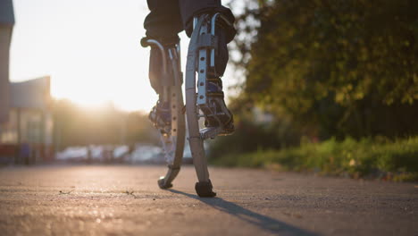 close-up view of legs walking on spring stilts in suburban outdoor setting with dark pants and sneakers. background features blurred trees, buildings, and a street. low camera angle highlights