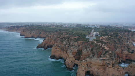 Amazing-aerial-drone-view-of-Lagos,-Portugal-rocky-coastline-with-lighthouse,-cloudy-grey-day,-circle-pan