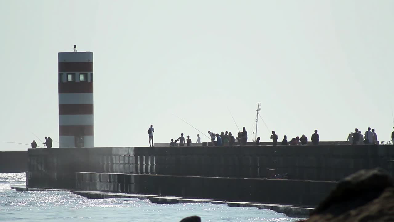 Silhouette of a lighthouse getting hit by big waves while tourists are walking along.