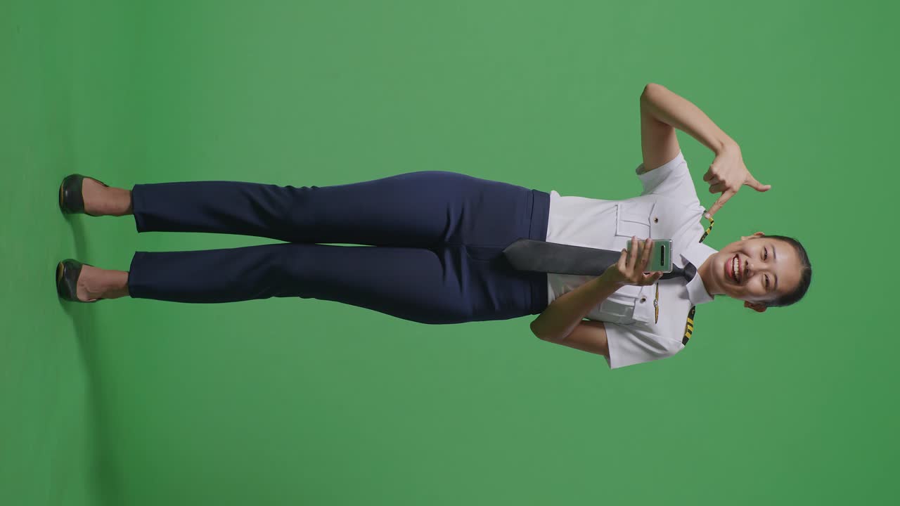 녹색 스크린 배경 스튜디오에 서 있는 동안 손에 스마트폰을 가리키며 미소 짓는 아시아 여성 조종사의 몸이 가득 차 있습니다.