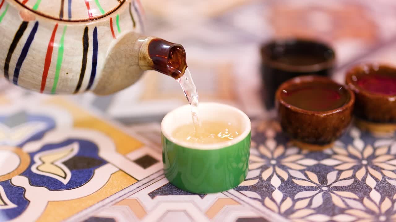 A teapot pours steaming tea into a cup on a vibrant, patterned tablecloth, creating a warm, inviting atmosphere