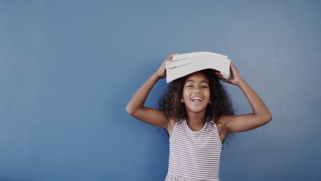 Retrato-De-Una-Chica-Afroamericana-Sosteniendo-Libros-En-La-Cabeza-Sobre-Fondo-Azul,-Espacio-Para-Copiar,-Cámara-Lenta