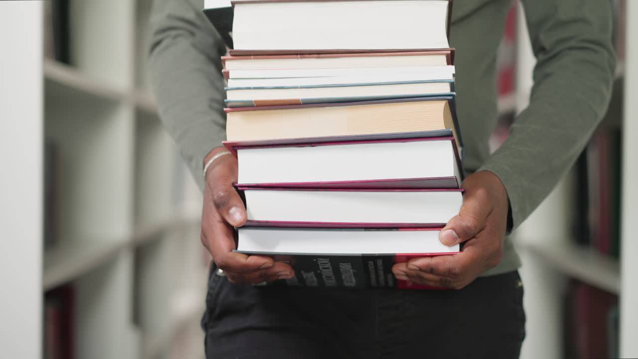 hombre negro lleva una pila de libros en una tienda de primer plano. maestra afroamericana sostiene una gran pila de libros de texto caminando entre estanterías. trabajo de bibliotecario