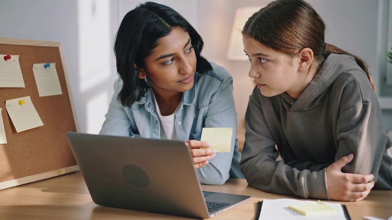 Picking up sticky note, denim tutor teaching grey hoodie student at desk with laptop for homework