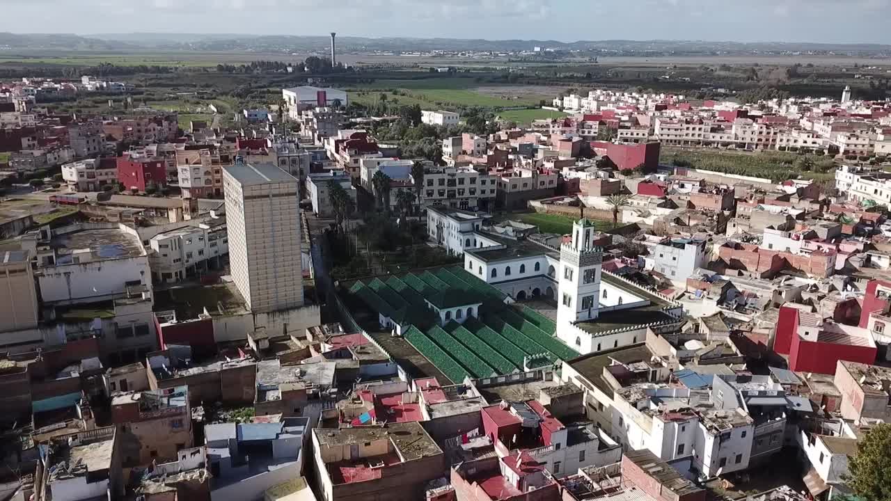 A detailed drone perspective of the historic Grand Mosque in Larache. Focuses on the elegant white minaret and the vast green-roofed prayer hall surrounded by the old city's urban texture