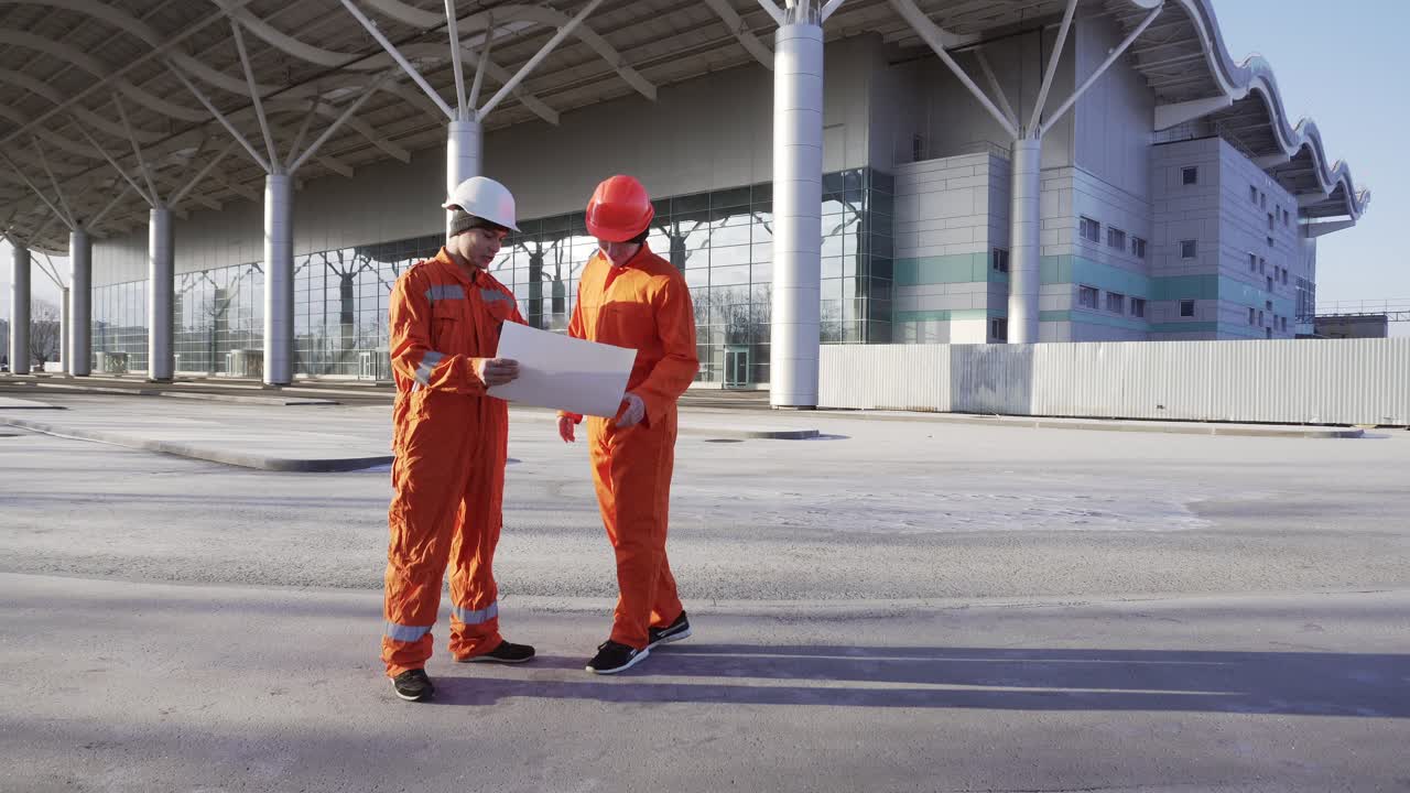 dos trabajadores de la construcción con uniforme naranja y cascos se reúnen en el objeto de construcción, estrechando la mano y examinando el edificio construido juntos. concepto de trabajo en equipo