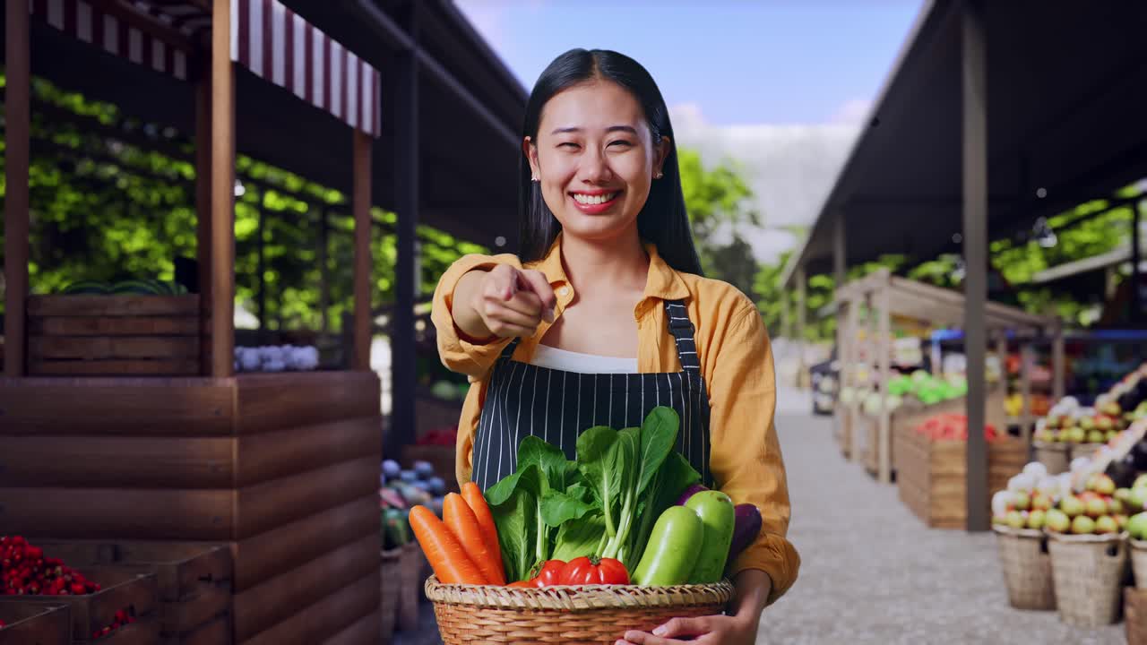 Agricultora asiática sonriente con una cesta de verduras haciendo un gesto de honestidad en un mercado de agricultores