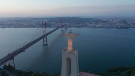 Aerial-evening-back-view-of-big-Jesus-statue-on-pedestal-and-long-bridge-over-river.-Drone-flying-around-Christ-the-King-Sanctuary-in-Almada-in-twilight-time.-Lisbon,-capital-of-Portugal.