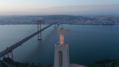 Vista-Aérea-Nocturna-De-La-Gran-Estatua-De-Jesús-En-Pedestal-Y-Largo-Puente-Sobre-El-Río.-Drone-Volando-Alrededor-Del-Santuario-De-Cristo-Rey-En-Almada-En-El-Crepúsculo.-Lisboa,-Capital-De-Portugal.