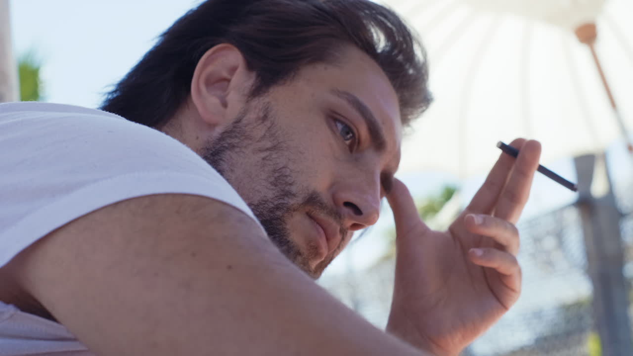 joven apuesto sentado al aire libre y sosteniendo un cigarrillo en la mano