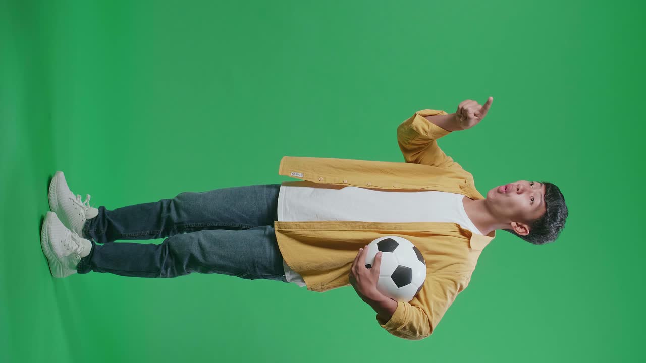 녹색 스크린 배경에서 축구를 응원하는 동안 패자를 조롱하는 공을 가리키는 아시아 소년의 전체 몸 측면 시각