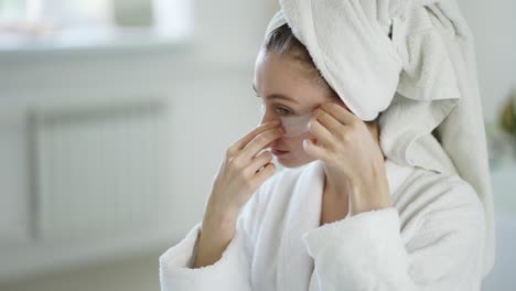 woman at home in a white bathrobe and towel applies patches under the eyes