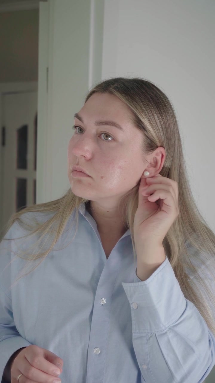 Giovane donna con una camicia blu si mette un orecchino davanti a uno specchio mentre finisce di prepararsi per un colloquio di lavoro online