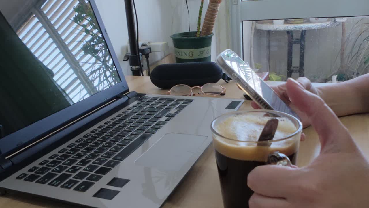 Scena di ufficio domestico: mani che usano lo smartphone accanto al laptop, caffè e accessori tecnologici
