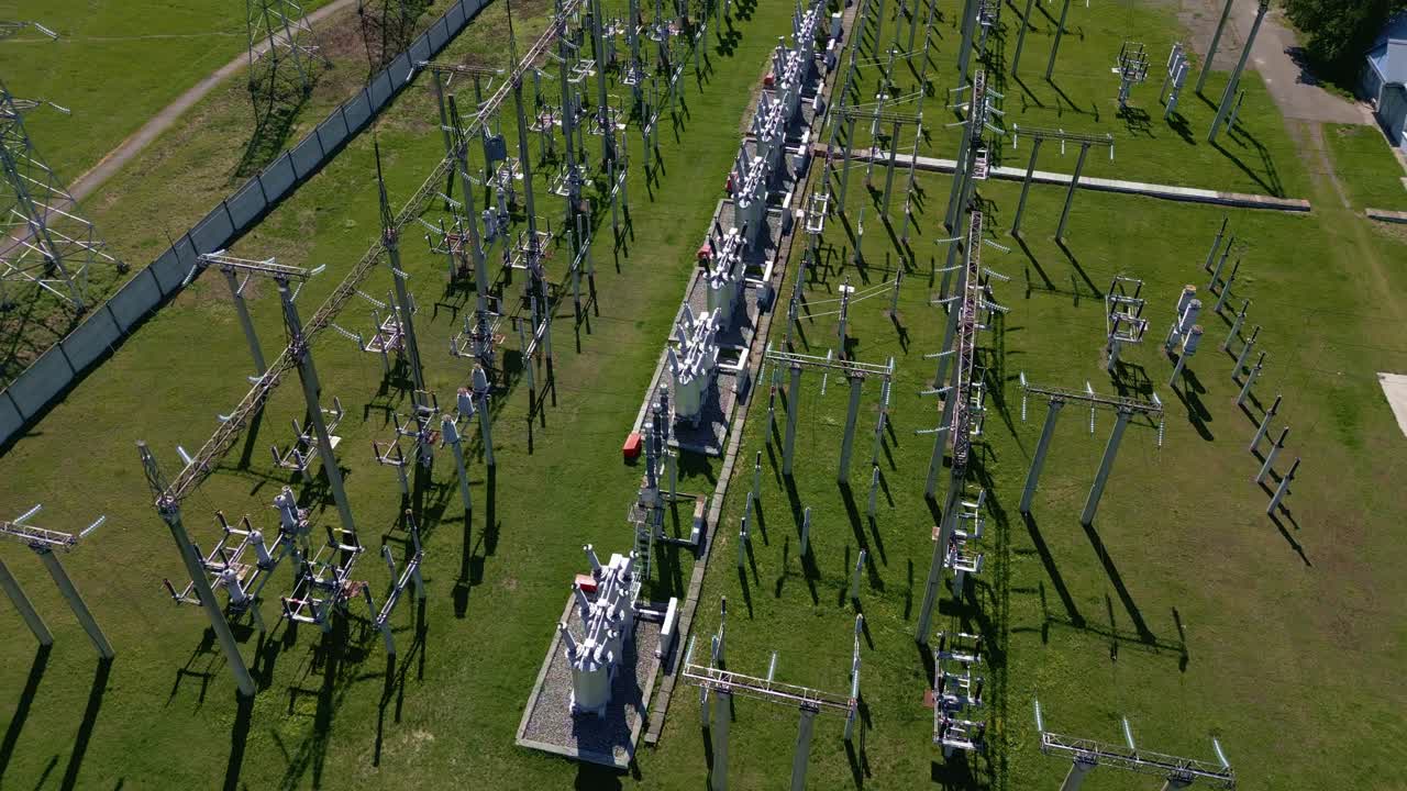 Letecký pohled ukazuje rozlehlou elektrickou rozvodnu. Kovové konstrukce lemují zelená travnatá pole. Dlouhé stíny se táhnou po zemi. Inženýři pracují u zařízení.