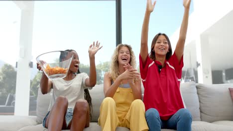 happy diverse female friends with chips watching sport on tv and cheering, slow motion