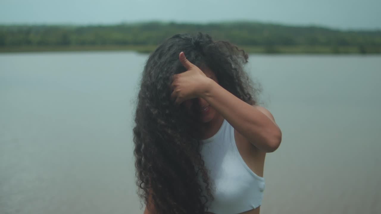 A young woman with voluminous dark curly hair and warm brown skin smiles joyfully as she tosses her hair with both hands near a calm lake, captured in slow motion under soft daylight