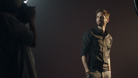 camera zooming in on the caucasian stylish young man posing to the camera of the female photographer in a dark studio during photosession