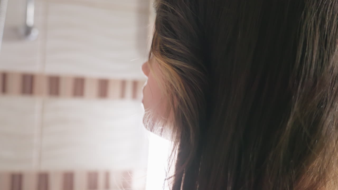 Back view young lady with long hair in well lit tiled bathroom stretches toward shelf to place item, soft highlights on hair and tiles, mirror and sink blur in background, everyday hygiene action