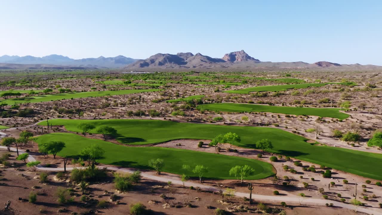Wide aerial view of a desert golf course surrounded by rugged terrain and mountain ranges under clear skies
