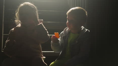 children eat crackers on stairs in evening. little boy and girl chewing biscuits silhouettes on staircase at rural house. kids rest at night camp