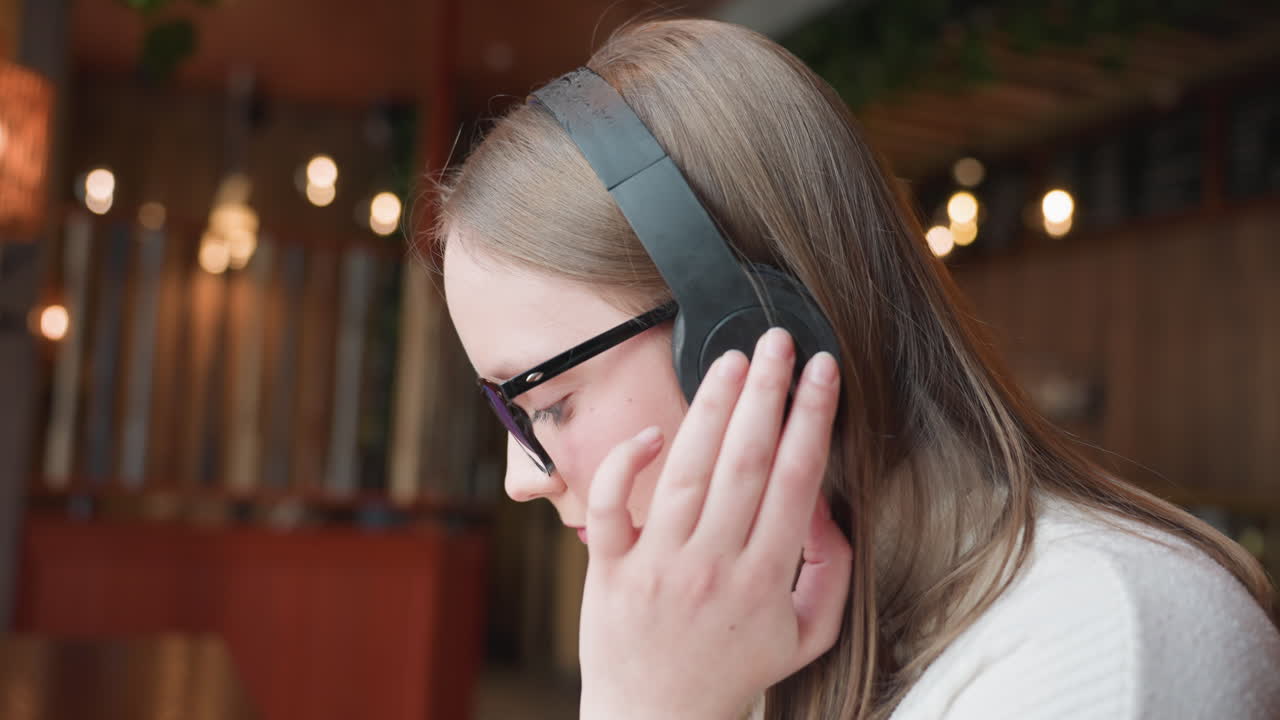 side view of young girl wearing black headphones and glasses with hand resting on ear as she dances joyfully in warmly lit indoor space with blurred golden lights in background