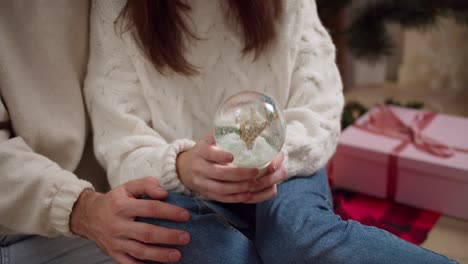 fotografía en primer plano de una chica morena con un suéter blanco sacudiendo una esfera de vidrio de año nuevo y su novio poniendo su mano en su regazo durante sus reuniones y comunicación en una acogedora habitación decorada de año novo