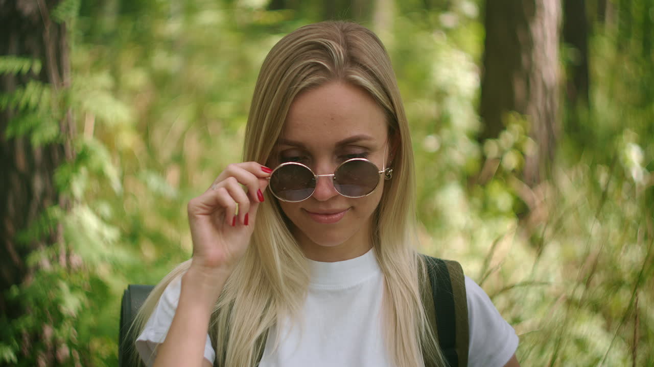 A young female traveler in sunglasses looks directly at the camera and smiles flirting a Traveler with a backpack in the Park and in the forest in slow motion