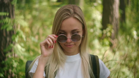 una joven viajera con gafas de sol mira directamente a la cámara y sonríe coqueteando con un viajero con una mochila en el parque y en el bosque en cámara lenta