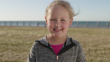 close up portrait of cute little blonde girl smiling happy enjoying sunny seaside park looking at camera shy