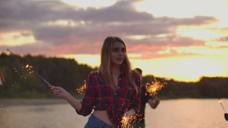 the girl with a mole on her chest is moving her hands with big bengal lights on the sand coast with her friends. this is theamy summer evening on the open air party.