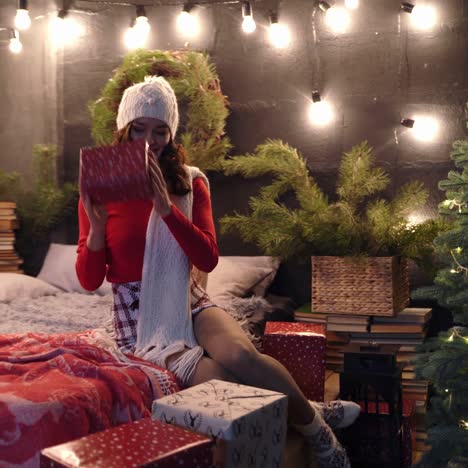 happy girl with gifts at christmas. attractive young woman in knitted white cap and scarf sitting on bed and looking at presents near new year tree at home.