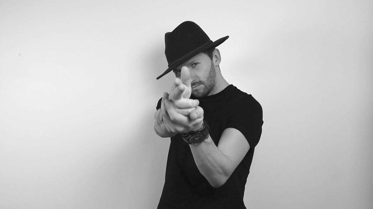 Filmato in scala di grigi di un uomo tatuato che indossa un cappello da cowboy o un elegante cappello rotondo, lo tocca e simula una pistola con le mani sparando alla telecamera. Successivamente soffia via il vapore e il fumo