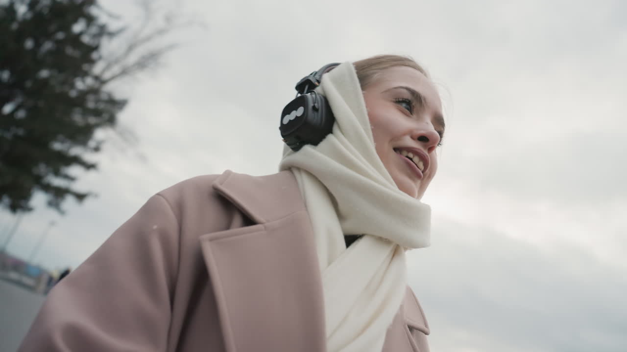 Jovem mulher contemplando o horizonte da cidade com fones de ouvido, cachecol envolvente e casaco de lã, expressões faciais tranquilas, respiração lenta sob céu nublado. Retrato urbano introspectivo com movimento suave e delicado.